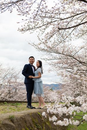 Couple in formal attire hold hands and pose beneath blooming cherry blossom branches during a spring engagement photo shoot outdoors.