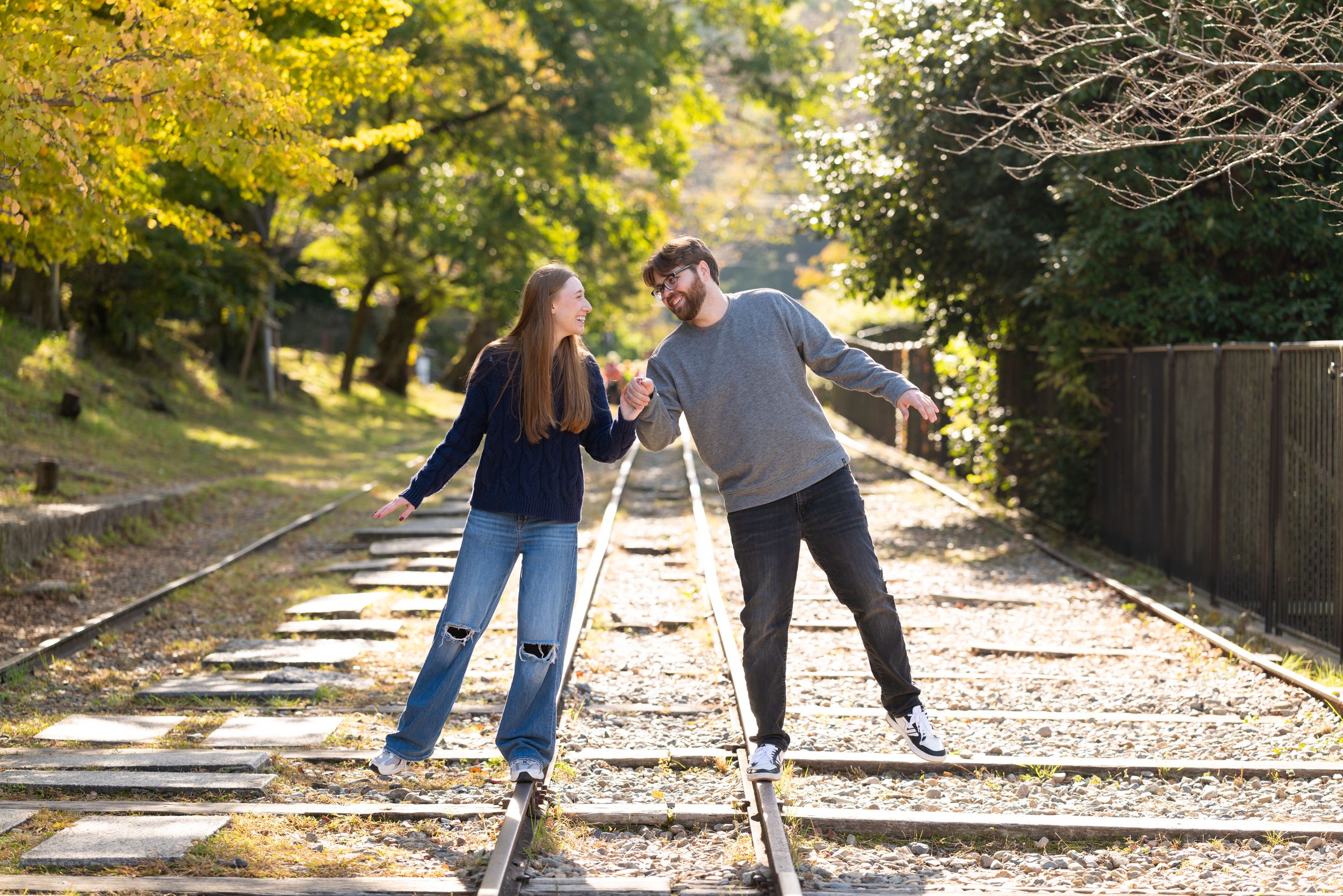 Smiling couple holding hands while balancing on old railway tracks surrounded by autumn trees on a sunny day.