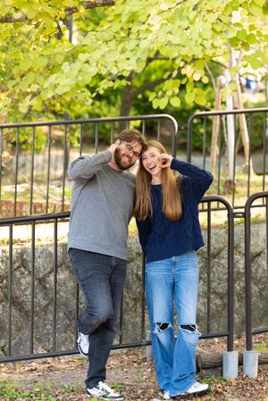 Smiling couple posing playfully with hands near their eyes beside a metal railing under green autumn leaves in a sunny park.