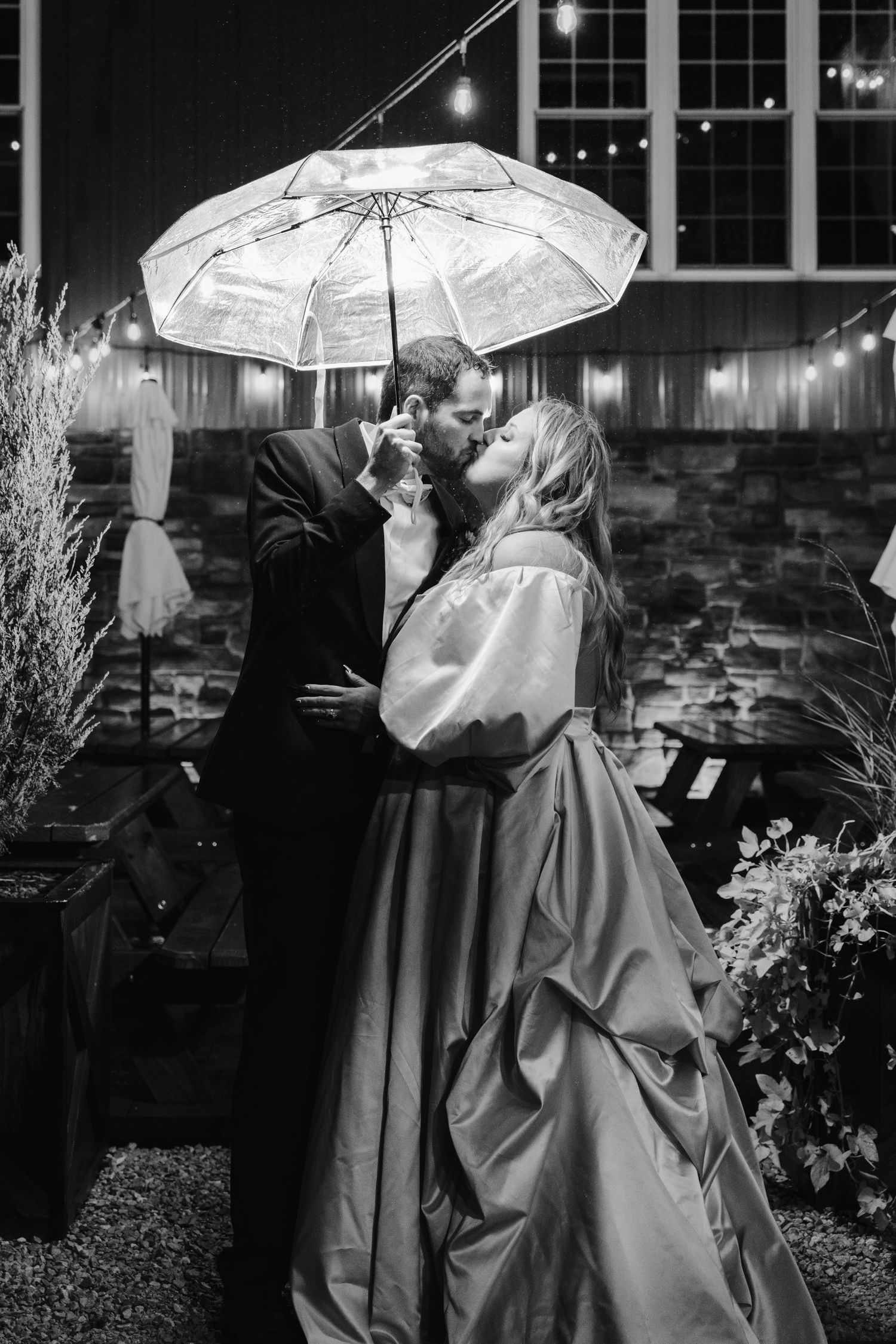 Romantic black and white photo of couple kissing under illuminated umbrella at night.