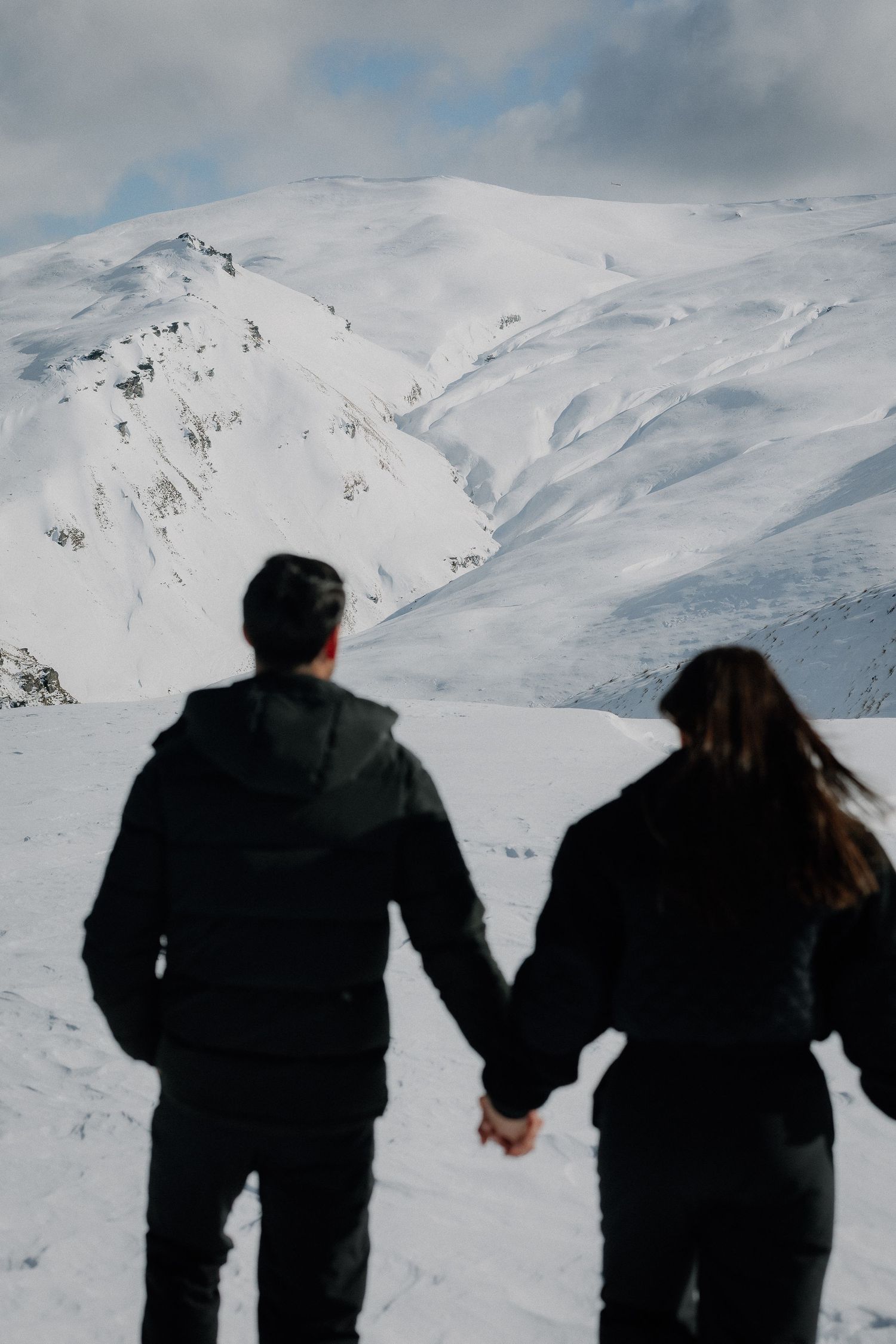 A hand with white nails and a diamond ring raised against a snowy mountain backdrop, wearing a black fur sleeve.