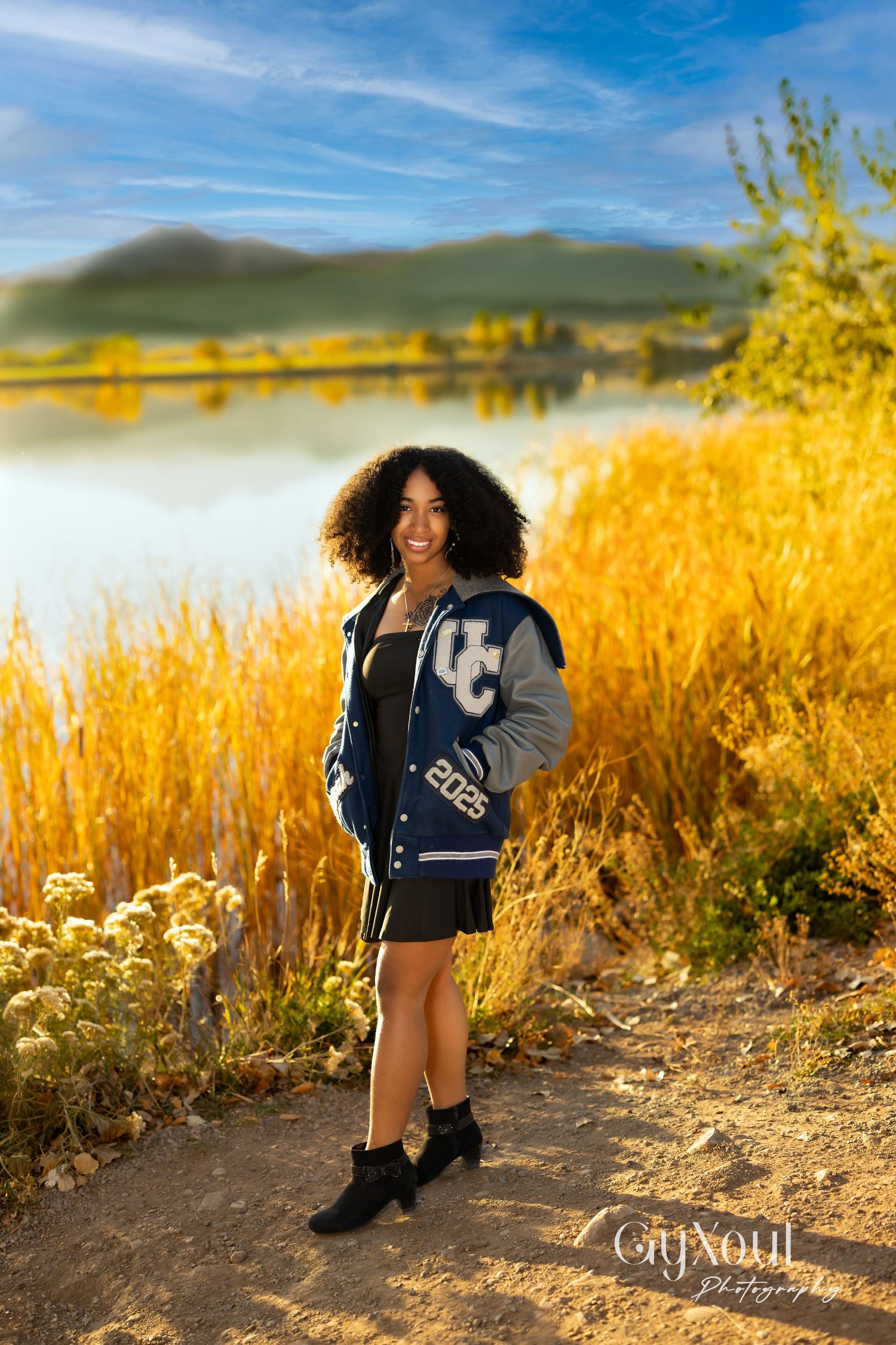 A smiling young woman in a UC 2025 varsity jacket poses on a trail by a reflective lake with golden autumn grasses.