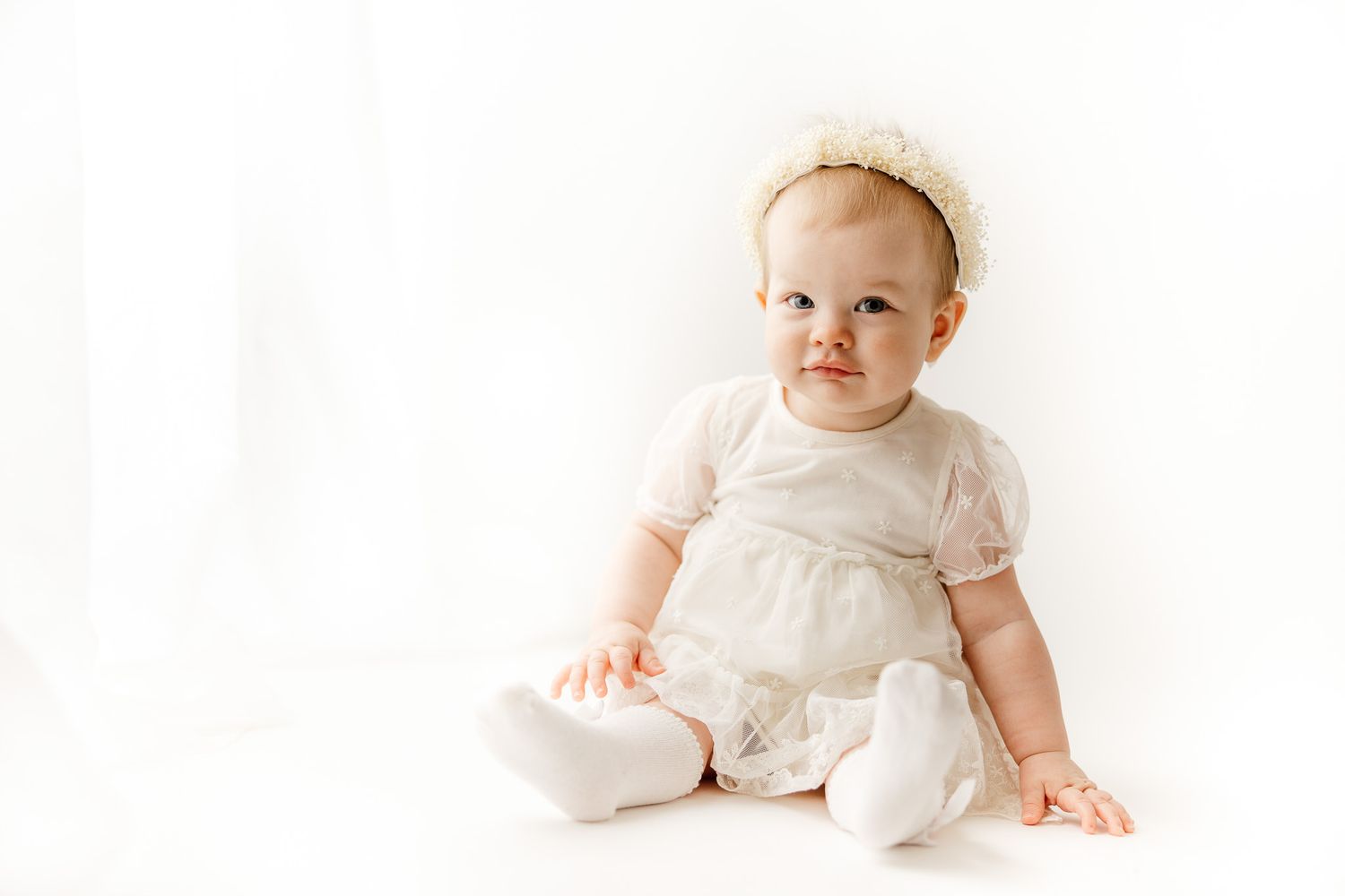 A baby girl in a white dress and headband sits on a white background, looking curiously to the side.