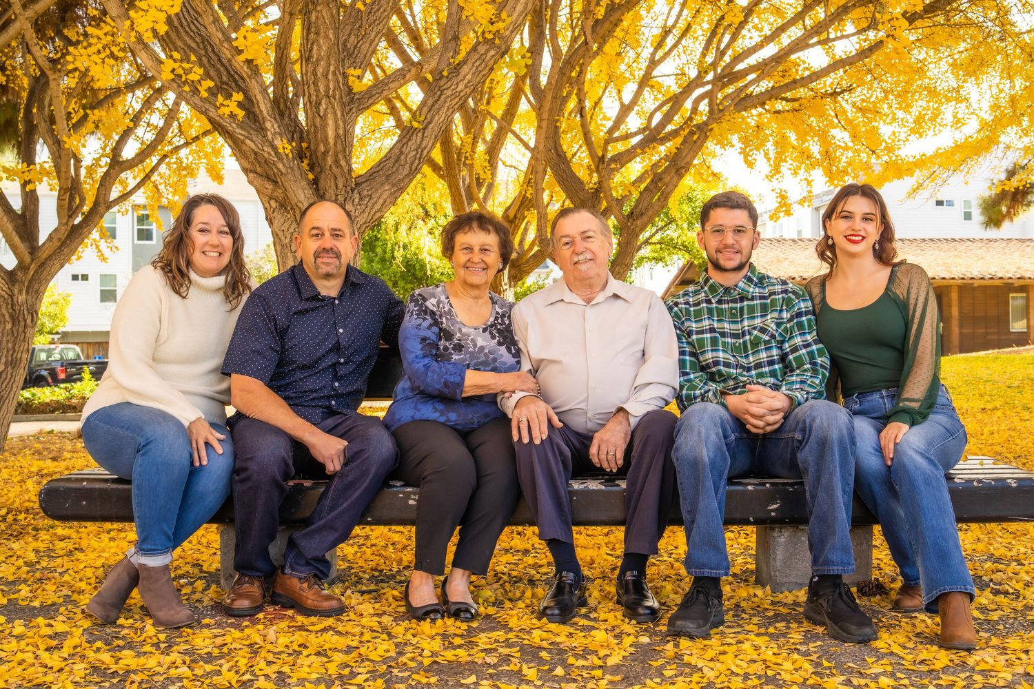 Family of six smiling together on a bench under golden autumn trees with yellow fallen leaves on the ground.