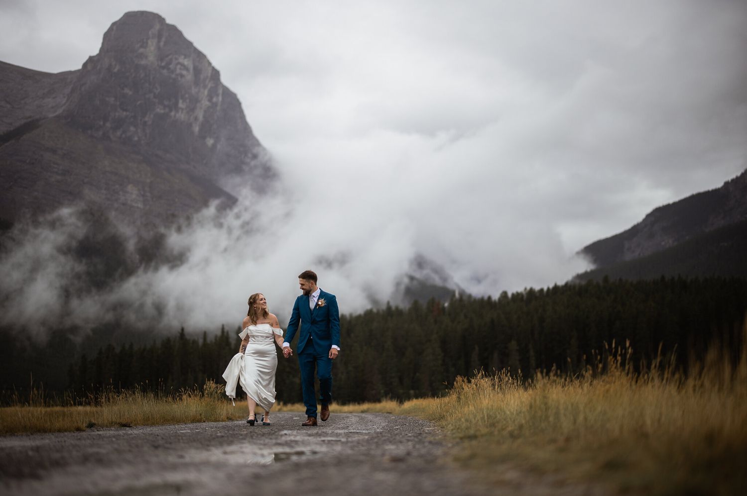 Mark and Jen walking hand in hand below misty peaks after their Canmore wedding