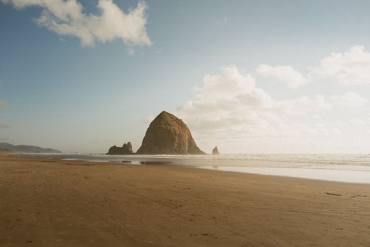 Sandy beach at low tide with iconic Haystack Rock rising from the ocean at Cannon Beach, Oregon under a partly cloudy sky.