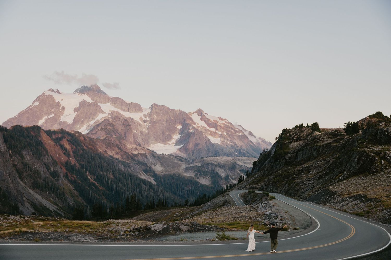 Bride and groom walking on a winding mountain road with snow-capped peaks in the background at dusk in Washington's North Cascades