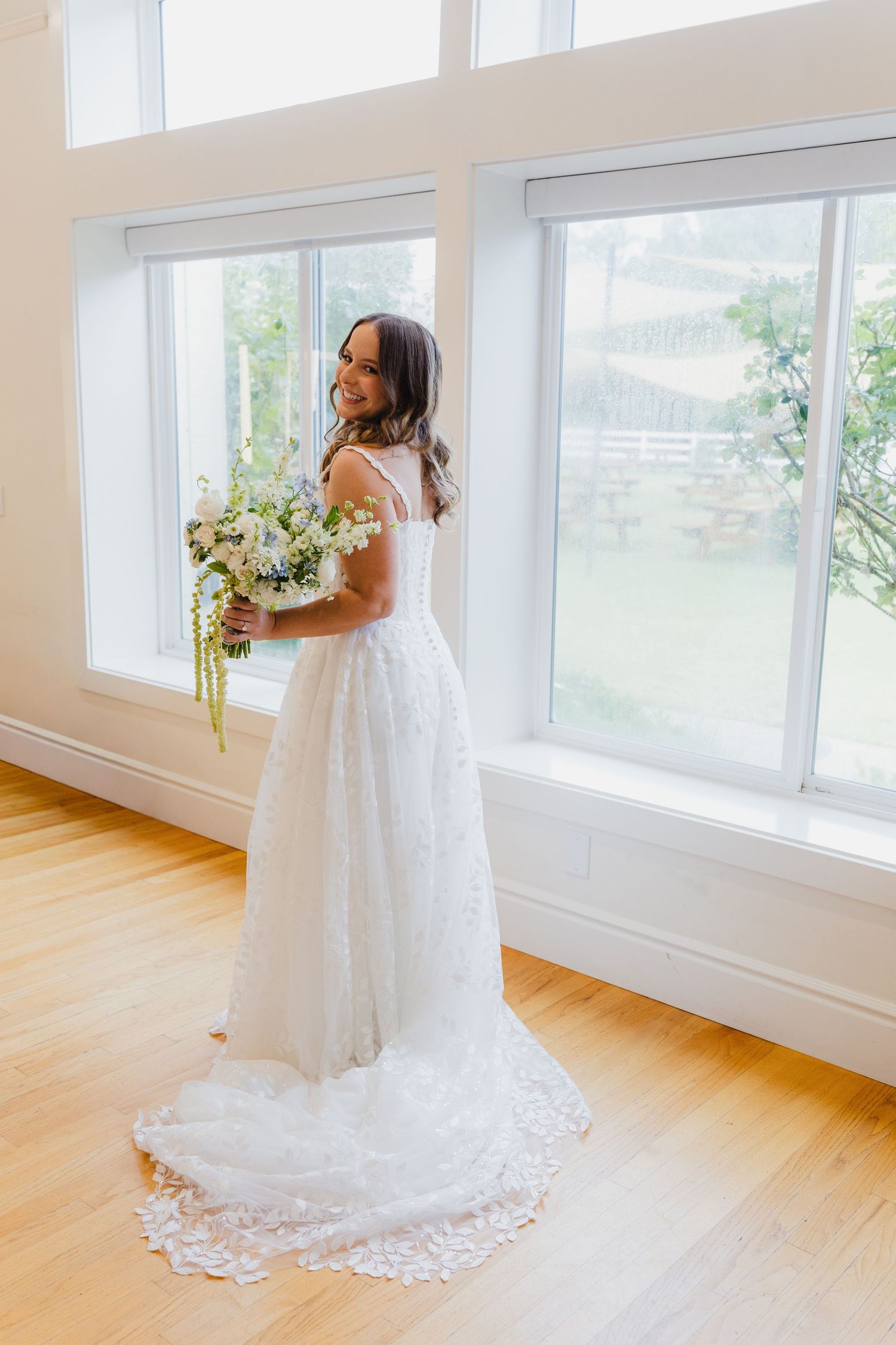 Smiling bride in a lace wedding dress holds a white and green bouquet while standing by large windows in a bright room.