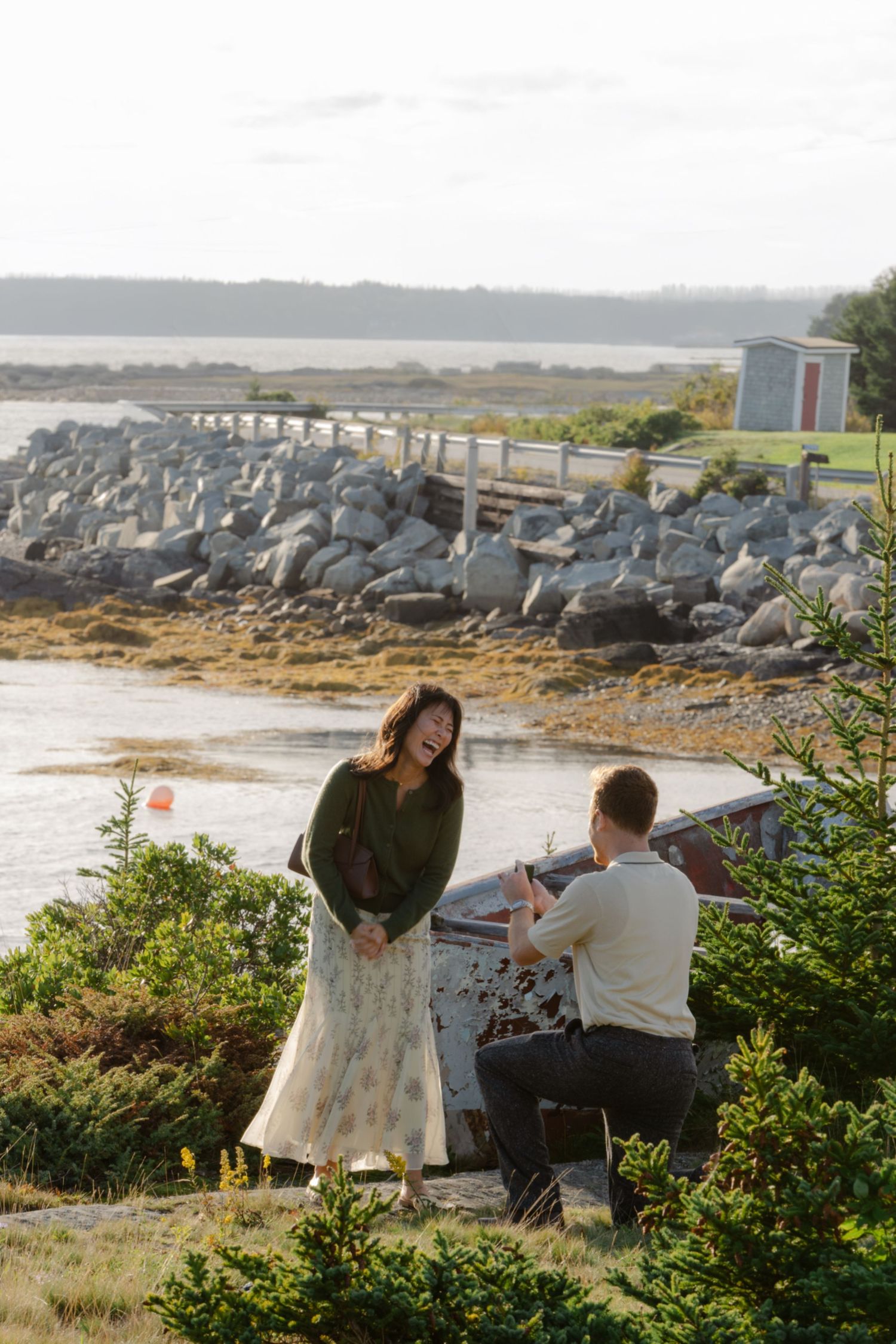 Man kneeling to propose to woman near rocky Nova Scotia coastline at golden hour.