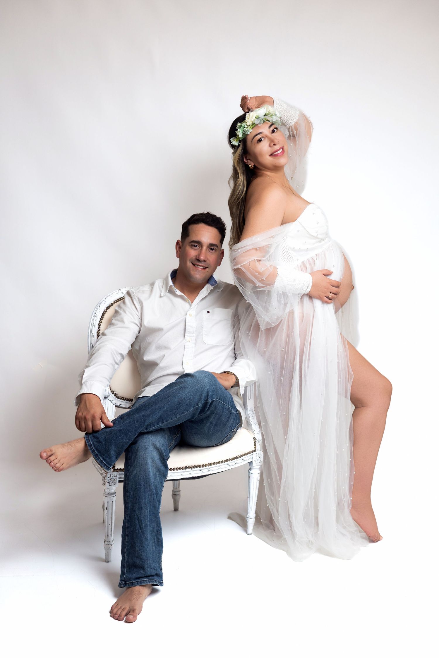 Pregnant woman in sheer white gown with floral crown poses with seated man in white shirt and jeans for maternity photo.