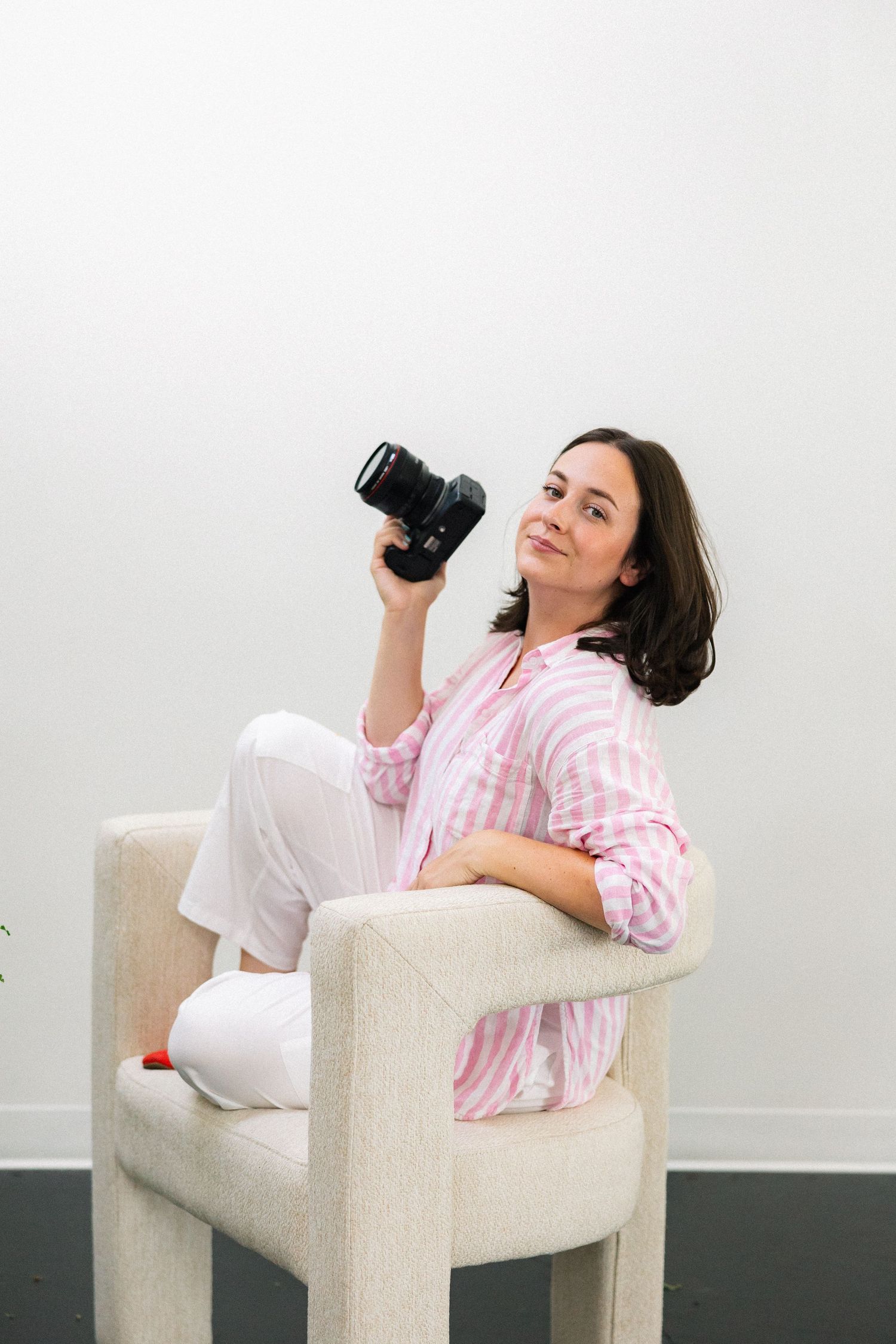 A smiling brunette woman in a pink striped shirt holds a DSLR camera while sitting relaxed in a modern white chair.