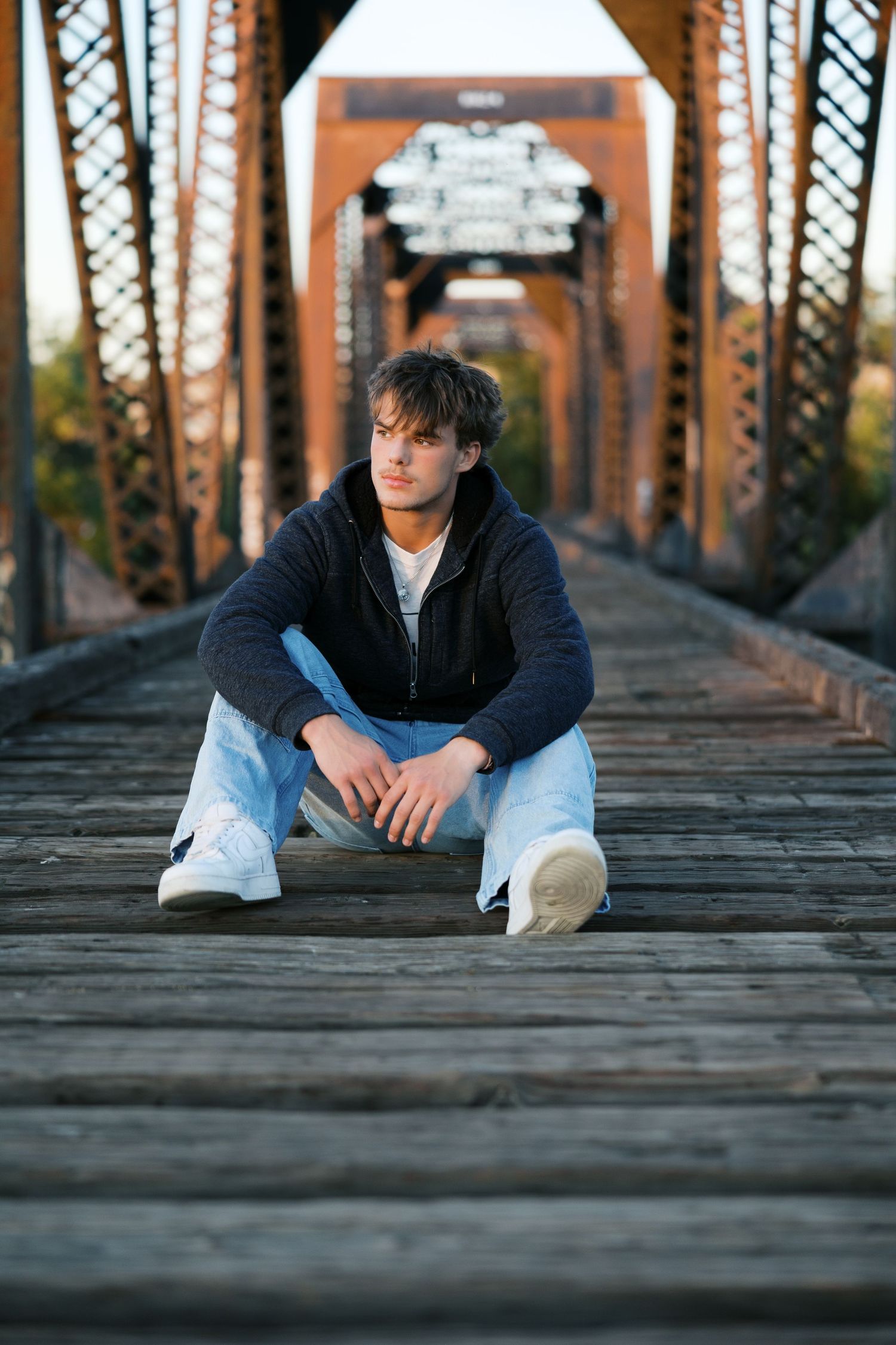 Young man in hoodie and jeans sitting on wooden bridge with rustic iron trusses in background.