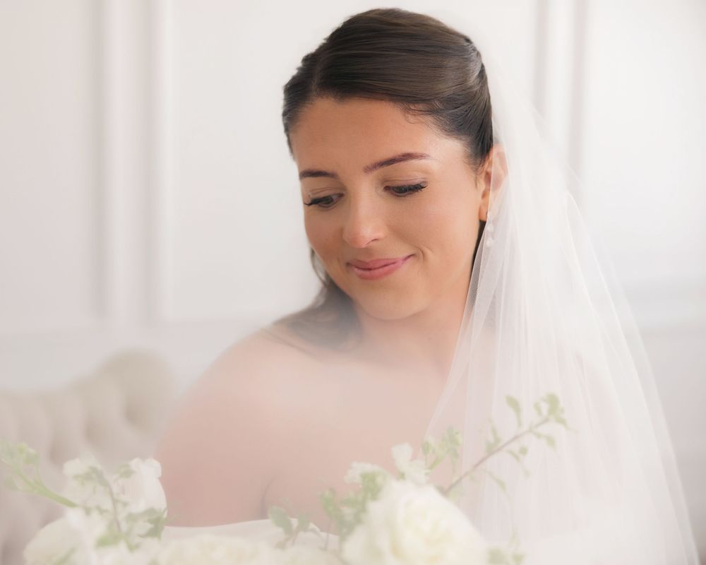 Bride in white veil looking down at bouquet of white flowers in soft natural light.