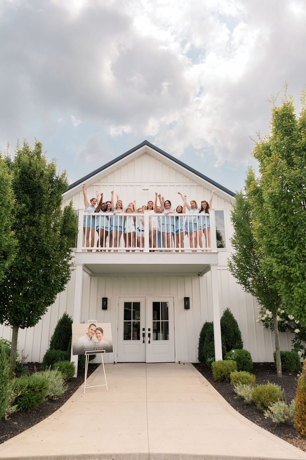 Group of women celebrating on a white barn venue balcony with trees and wedding photo display at entrance.