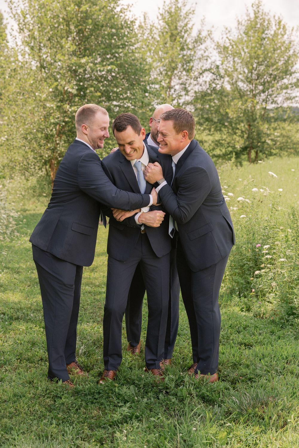Three groomsmen in navy suits laughing and embracing outdoors on a green field at a wedding.