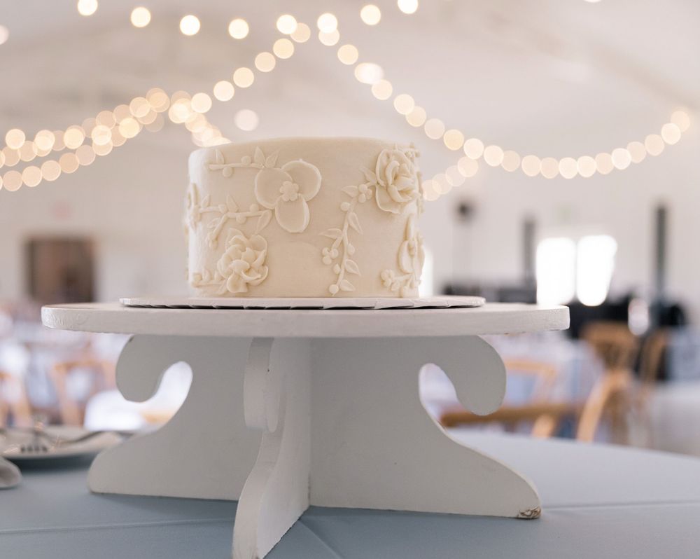White wedding cake with floral details on a white pedestal stand, surrounded by soft bokeh string lights.