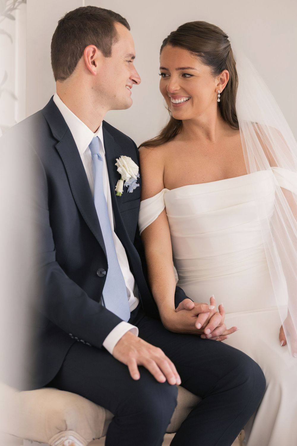 A bride and groom sit together smiling, she in a white off-shoulder gown with veil, he in a navy suit with light blue tie.