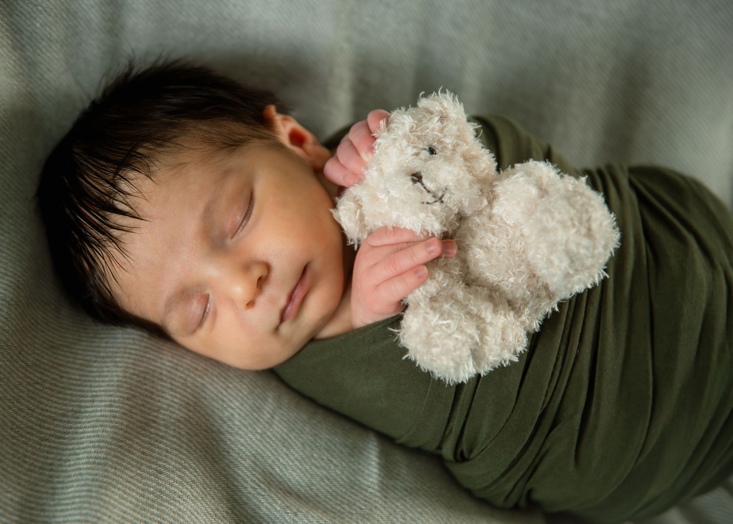 Sleeping newborn baby swaddled in green wrap holding a small white teddy bear stuffed animal.