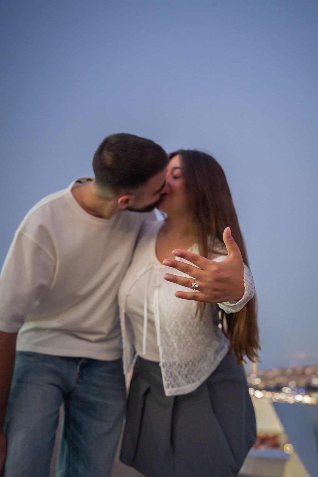 Couple sharing a kiss at dusk while woman shows off her engagement ring with city lights in background.