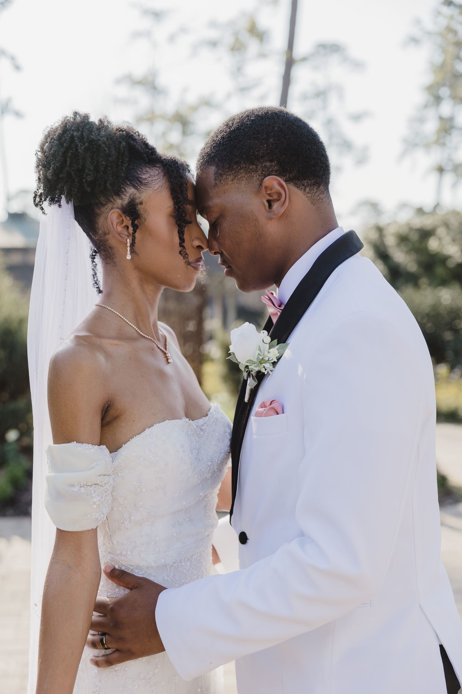A Black bride and groom in white wedding attire share an intimate forehead touch outdoors on their wedding day.