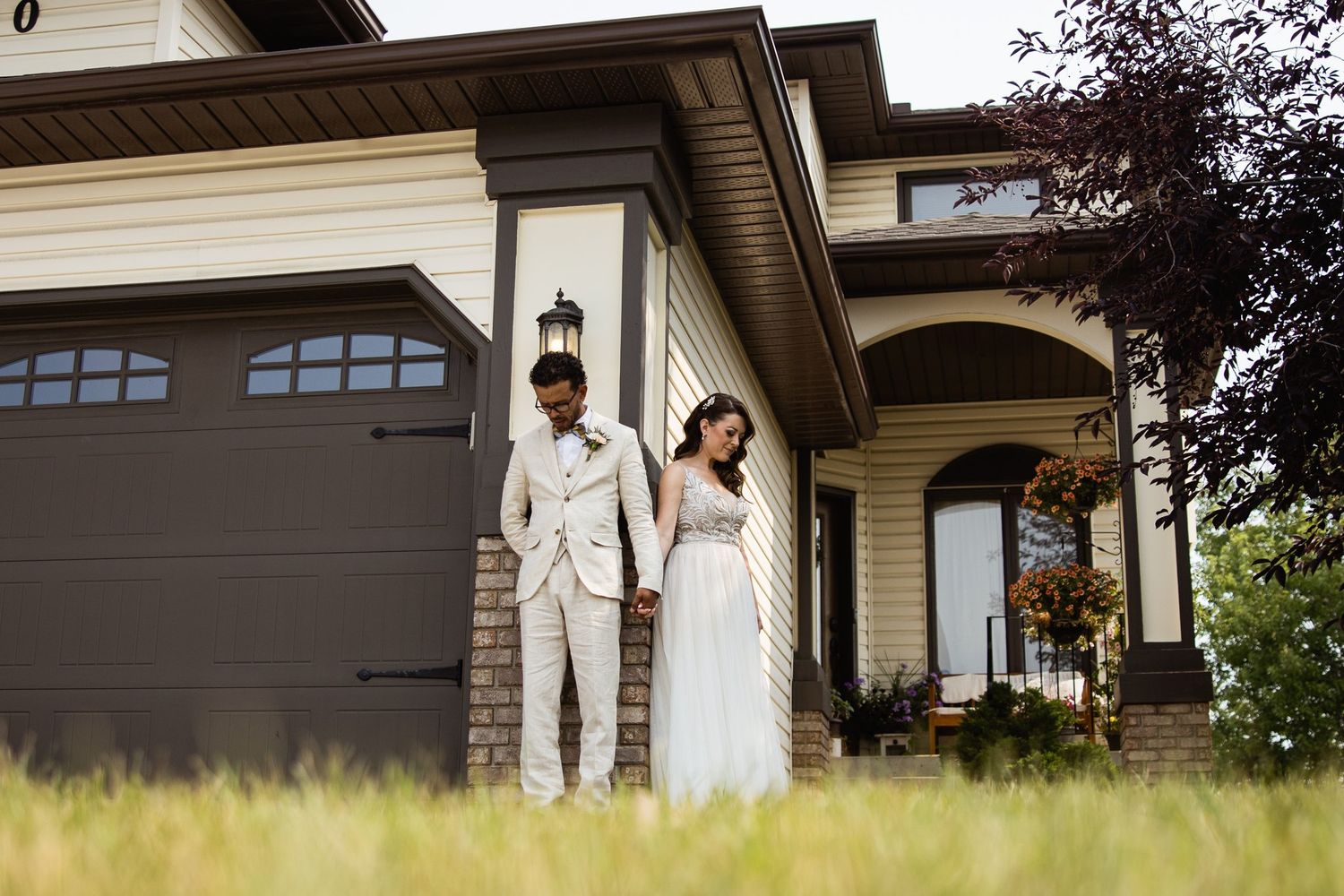 Wide low view from the grass of the bride and groom at the brick garage pillar, heads bowed in prayer and holding hands