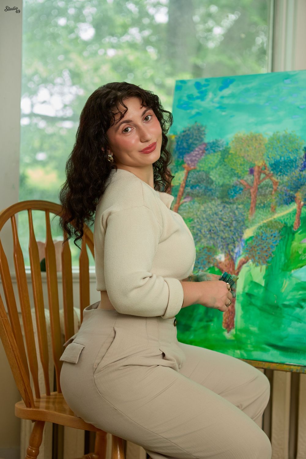 A young woman with curly dark hair sits on a wooden chair beside a vibrant green landscape painting in a bright studio.