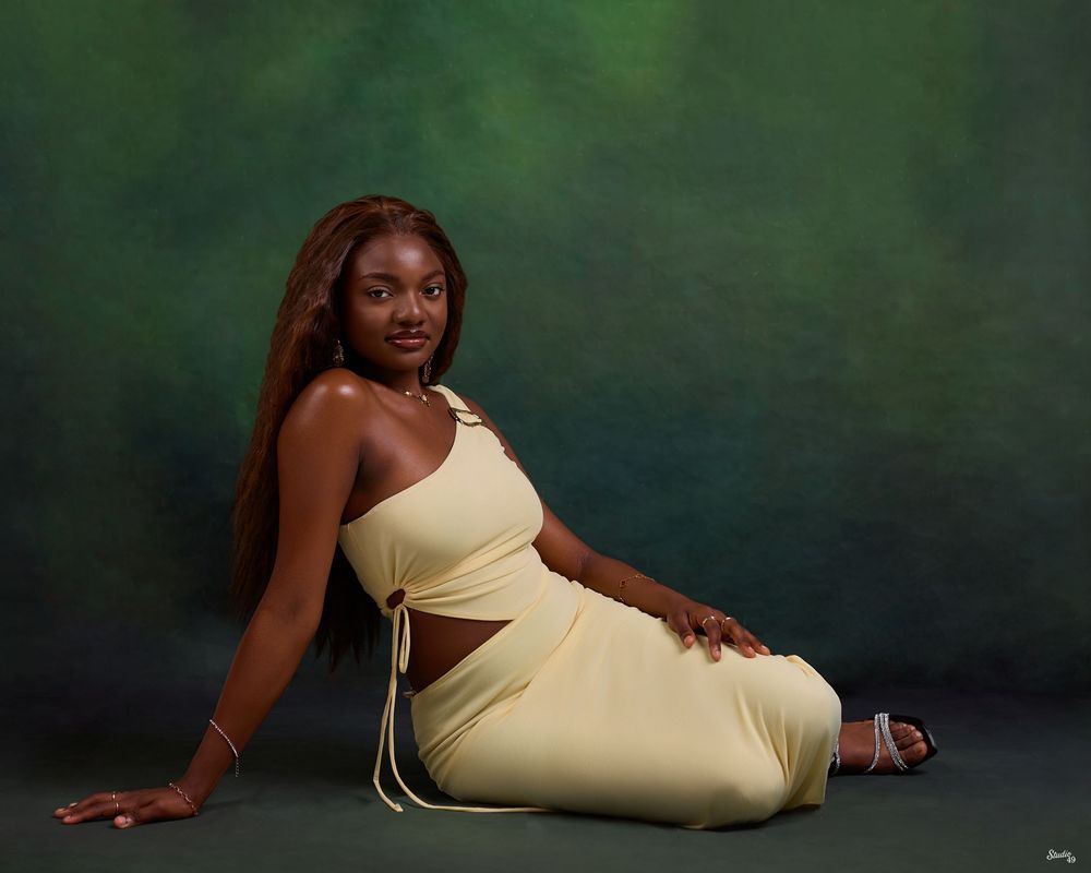 Young woman in yellow crop top and skirt set poses gracefully on floor against green backdrop.