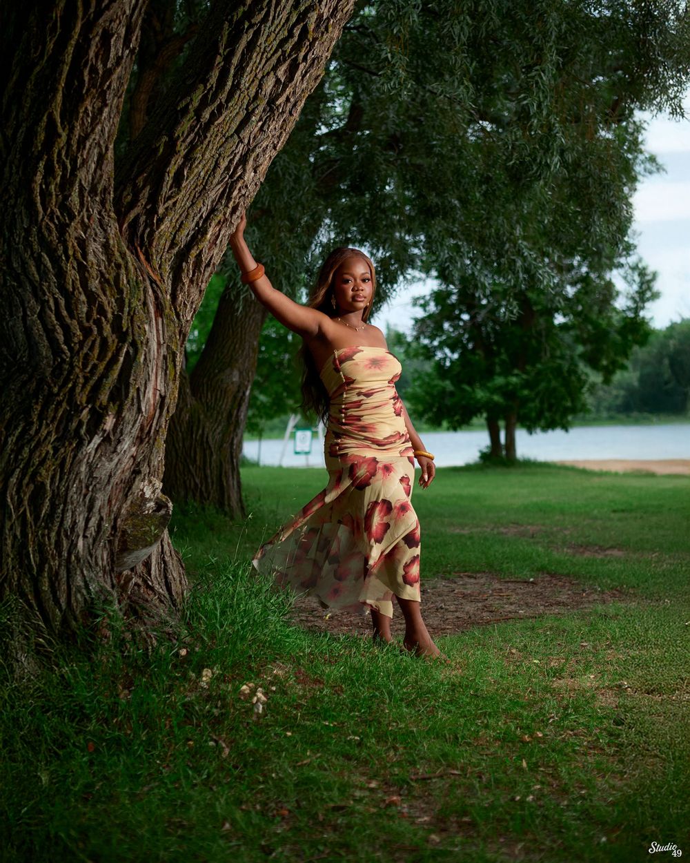 Woman in floral dress posing gracefully by a large tree in a lush green park near a lake.