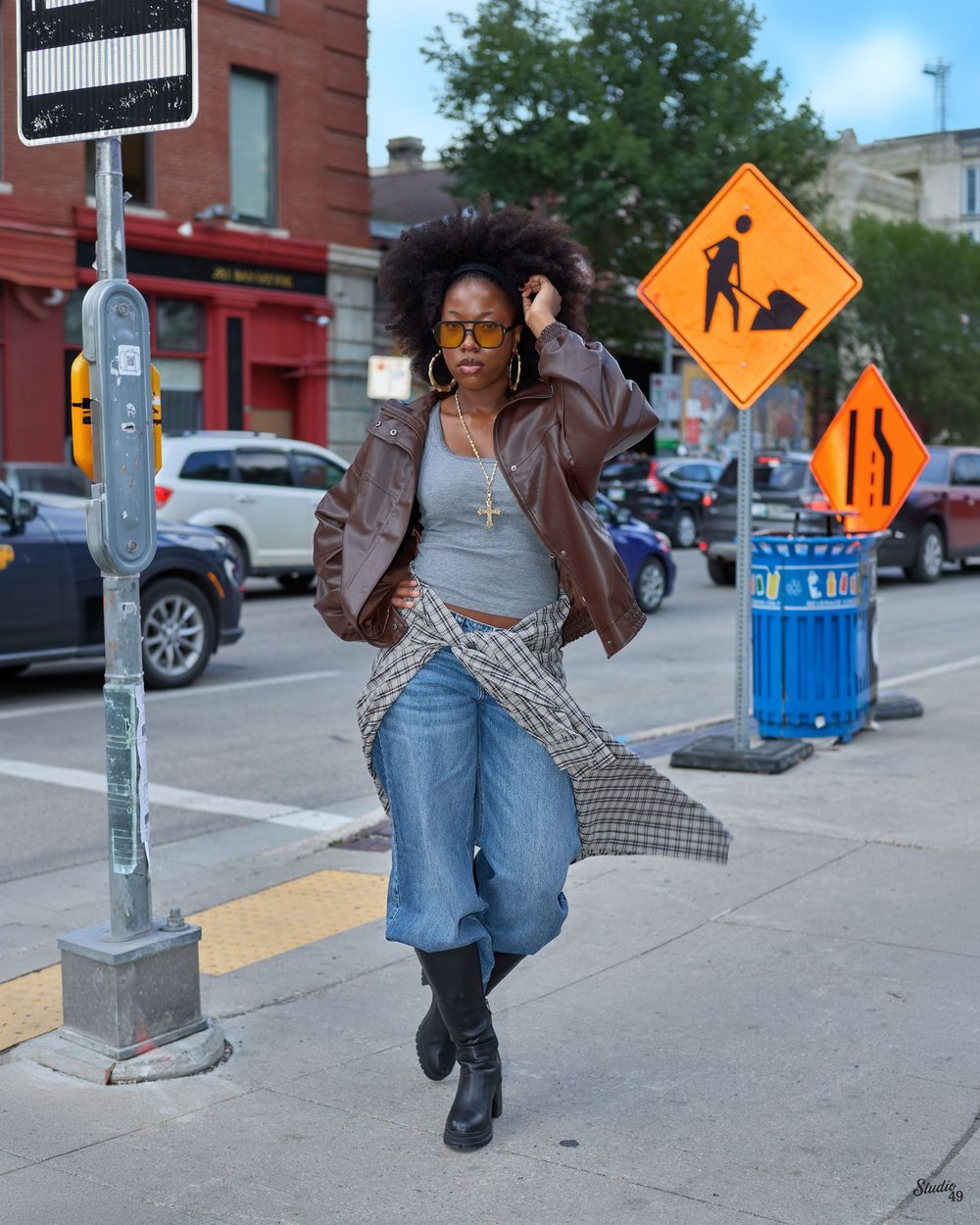Stylish woman in brown leather jacket, jeans, and boots poses on urban sidewalk near construction signs.