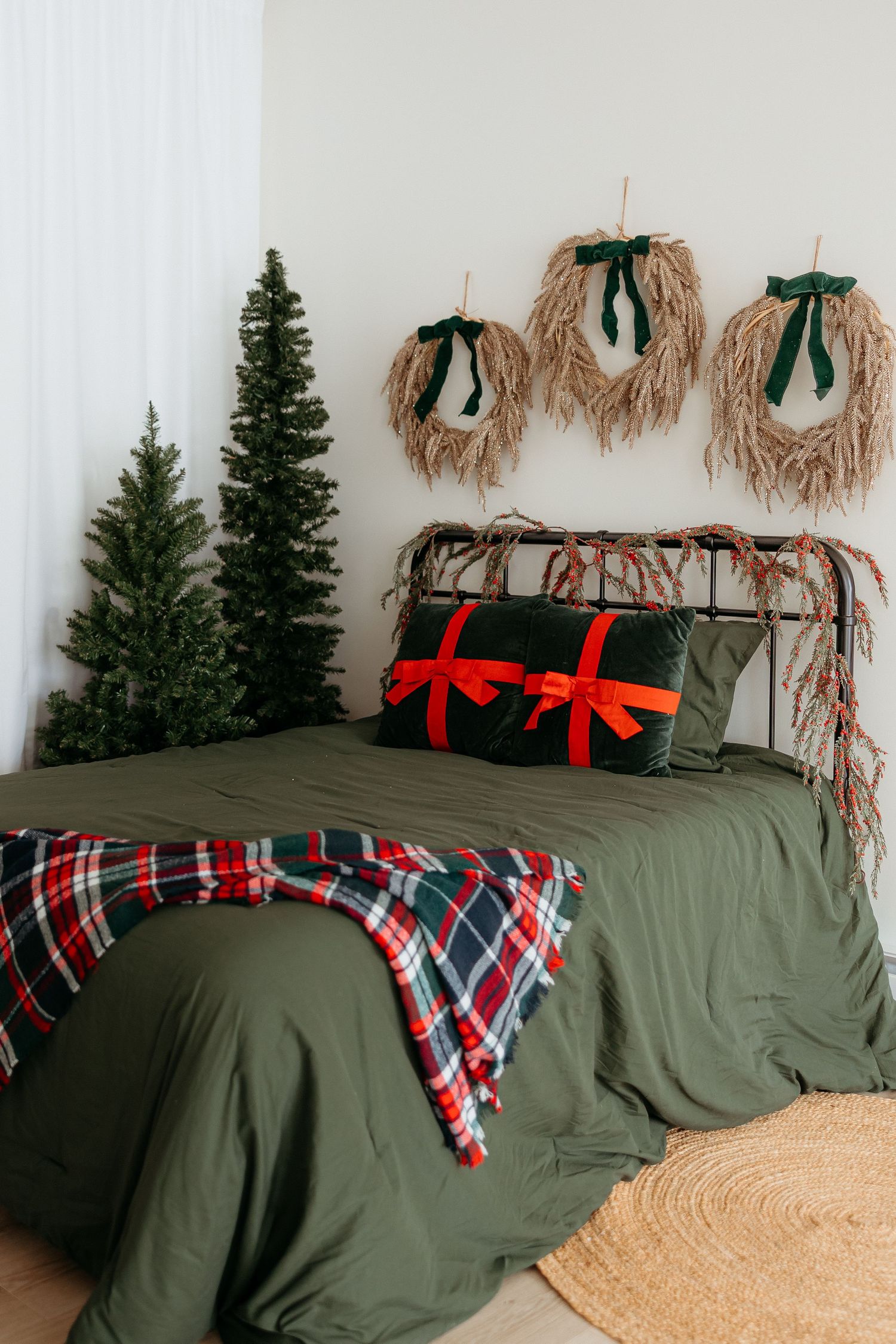 Christmas bedroom with green bedding, plaid throw, gift bow pillows, mini trees, and rustic wreath wall decor.