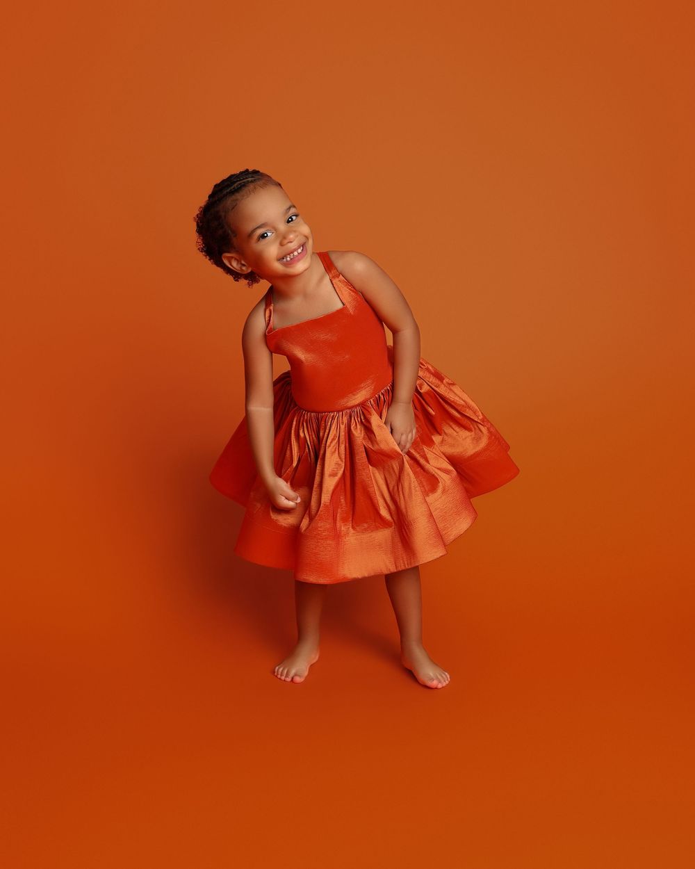 Young girl in orange tiered satin dress poses barefoot against matching orange background, smiling joyfully.