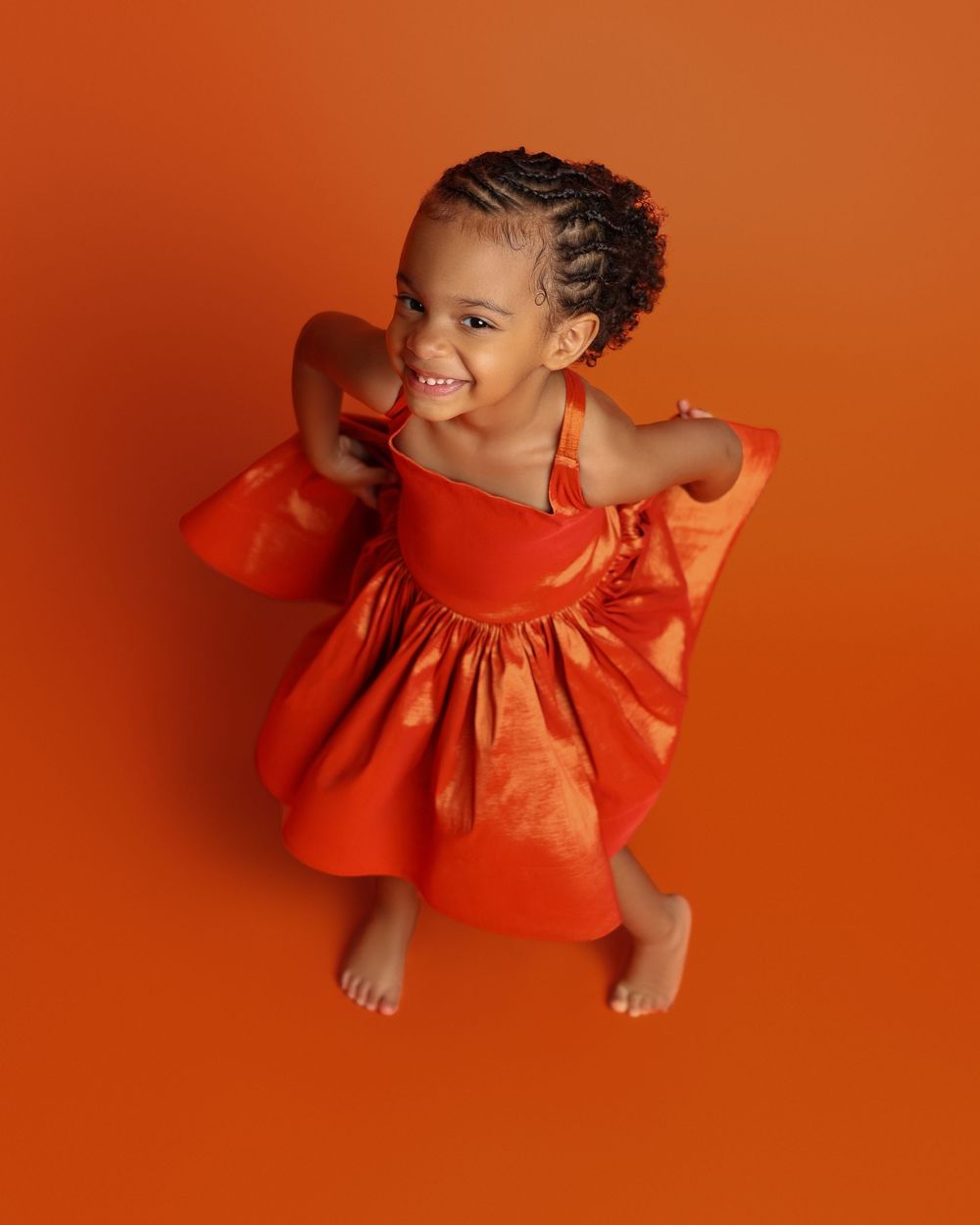 A joyful young girl in an orange dress poses barefoot against a vibrant orange background, smiling brightly.