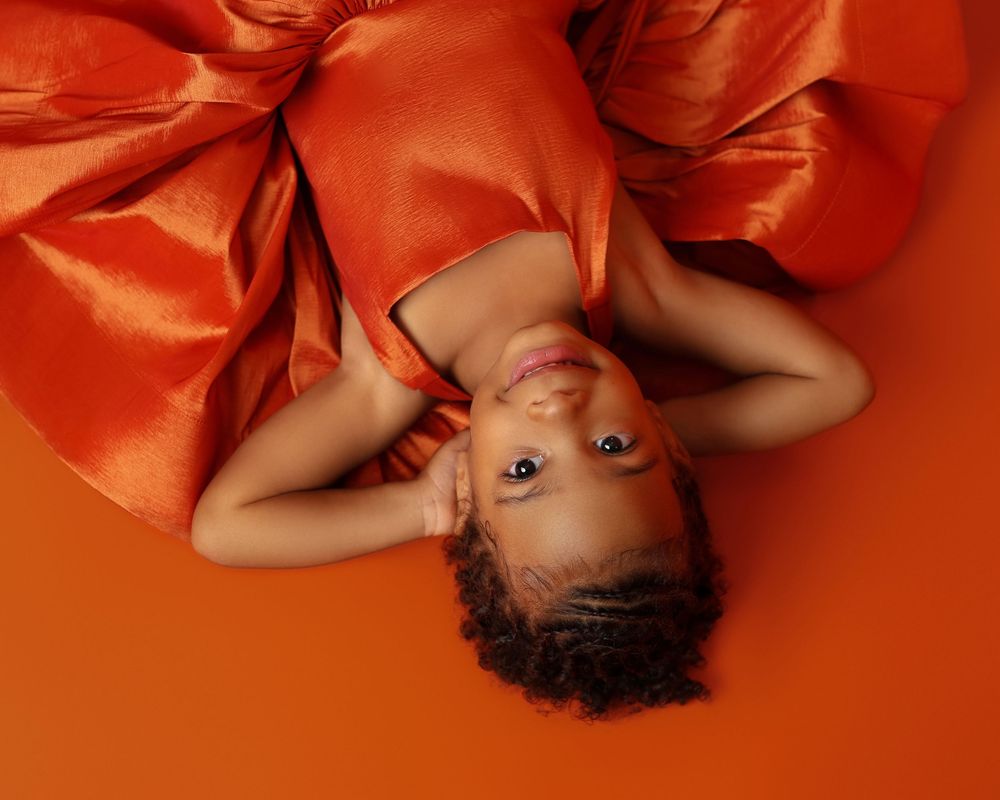 Toddler girl in orange silk dress lying on orange background, looking up at camera with natural curly hair.