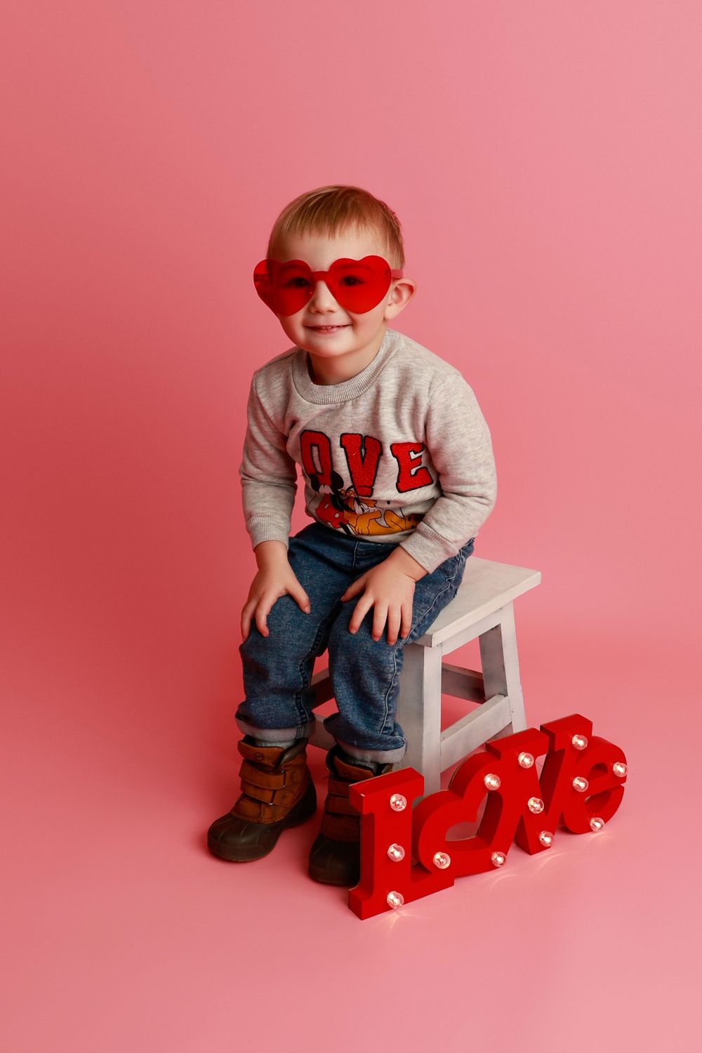 Cute toddler boy wearing heart-shaped sunglasses and LOVE sweater sitting on white stool with red LOVE letters on pink background in Covingt