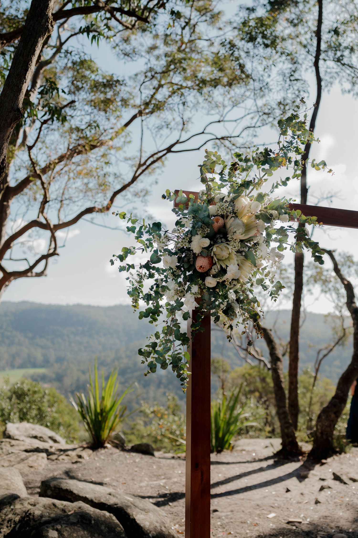 Rustic wooden cross adorned with white and blush floral arrangements overlooking a scenic mountain valley at an outdoor wedding.