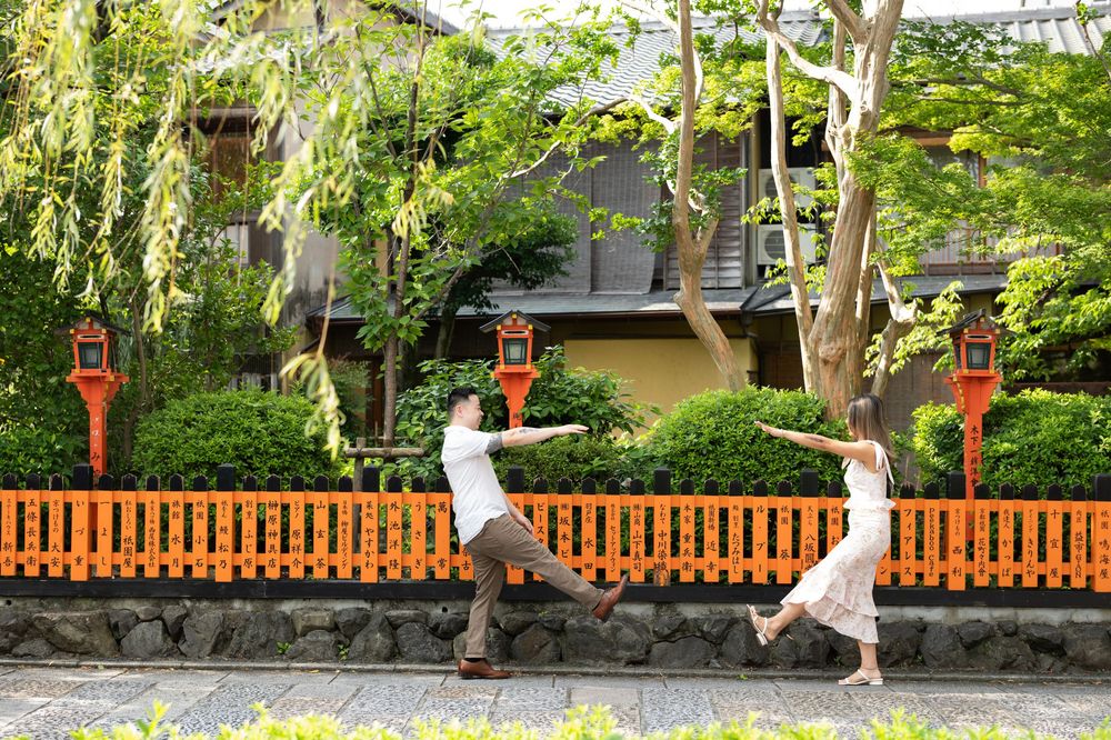 Couple in white playfully posing with outstretched arms in front of an orange wooden fence and lanterns in a lush green Japanese garden.