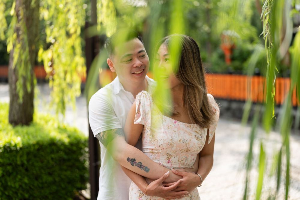 Smiling couple embracing in a lush green garden, the man in a white shirt and the woman in a floral dress, framed by willow branches.