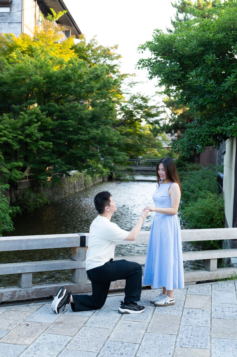 Man kneeling on one knee proposing to a woman in a blue dress on a stone bridge beside a calm river surrounded by lush green trees.
