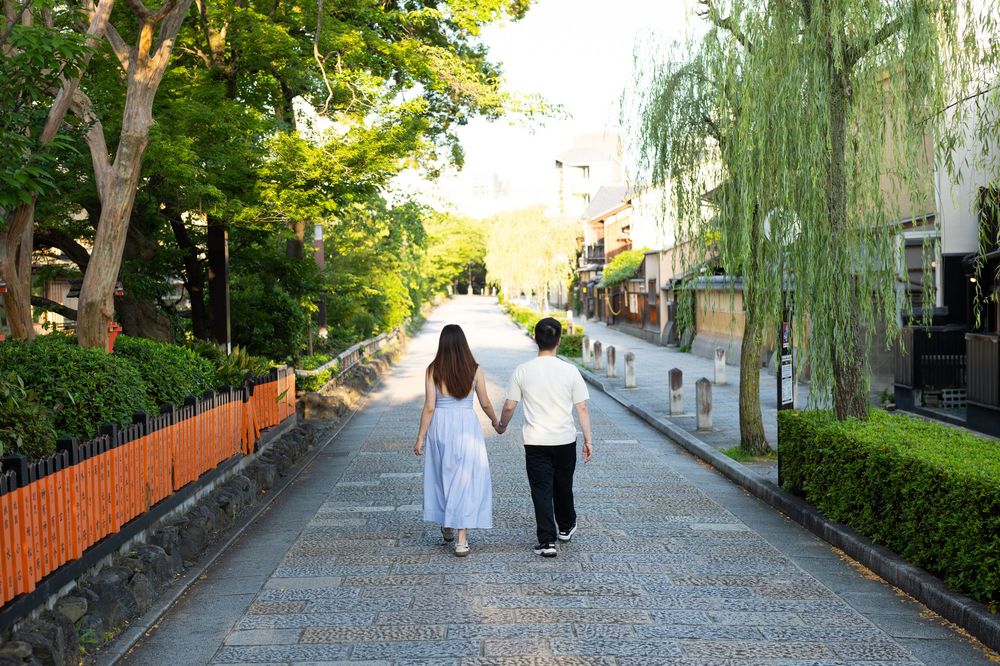 Couple holding hands walking along a quiet stone-paved path lined with green trees, weeping willows and an orange wooden fence.