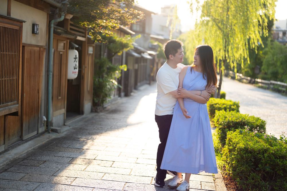 Couple embracing on a quiet stone-paved lane lined with traditional wooden buildings and willow trees in warm golden sunlight.