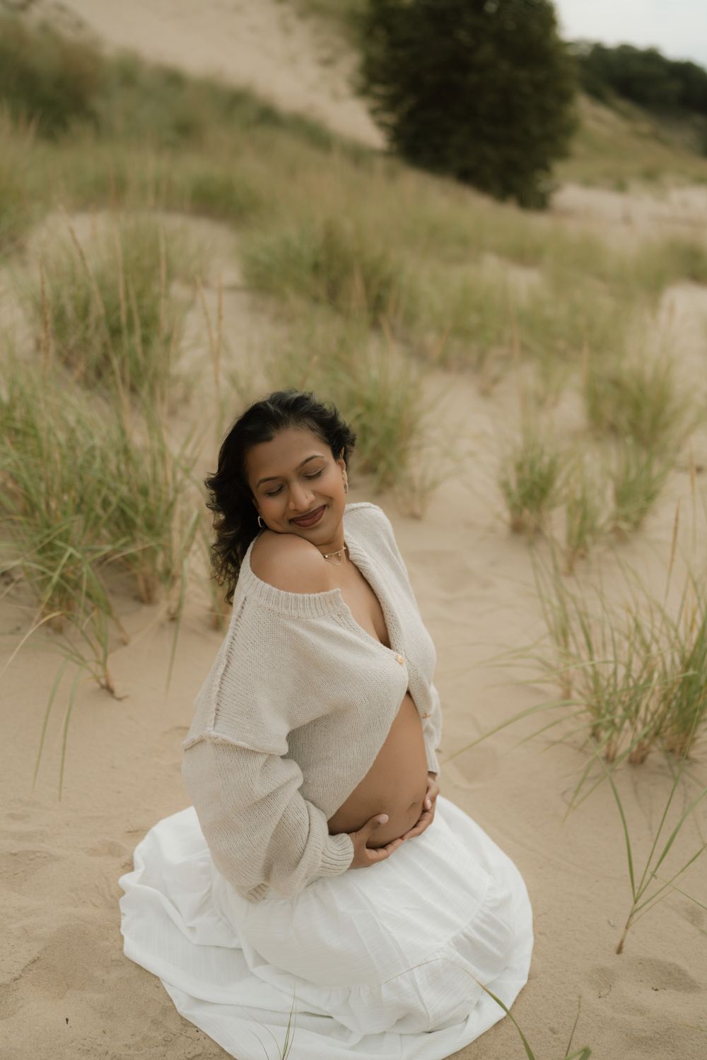 Smiling pregnant woman in a cream off-shoulder sweater and white skirt sits on a sandy dune, cradling her baby bump among beach grass.