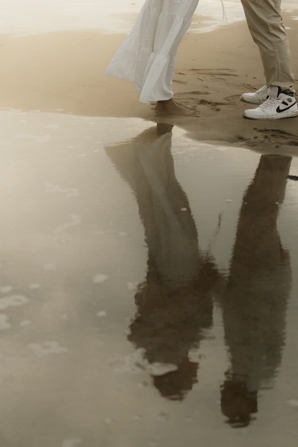 Two people walking along a wet sandy beach, their legs and white sneakers reflected in the shallow water in soft, muted tones.