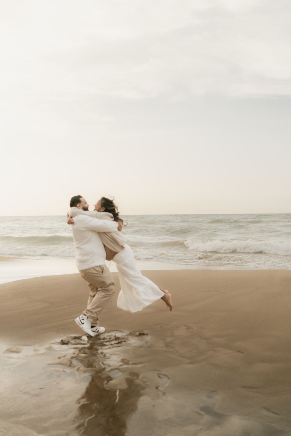 Couple embracing as the man lifts the woman in a white dress on a wet sandy beach with ocean waves at sunset.