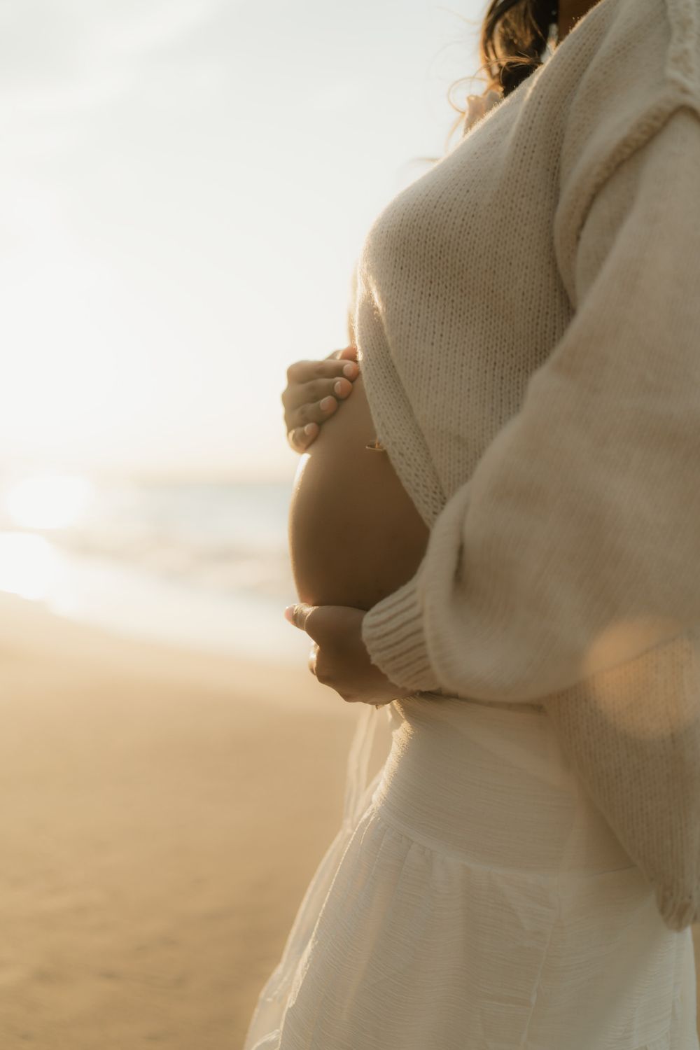 Pregnant woman in a cream knit sweater and white skirt cradles her bare belly on a sunlit beach at golden hour.