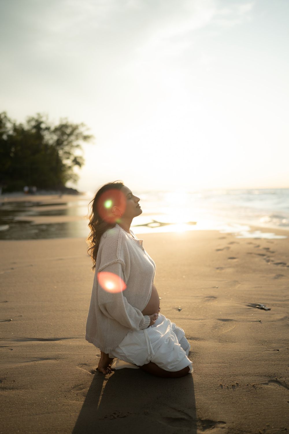 Woman in a white linen outfit kneeling on a sandy beach at golden hour, face tilted toward the glowing sun with soft lens flare.