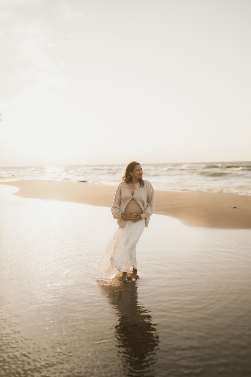 Pregnant woman in a white skirt and beige cardigan cradles her belly while standing in shallow water on a beach at golden sunset.