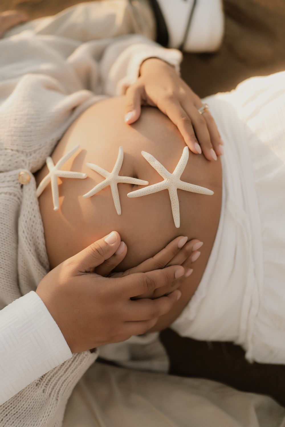 Pregnant woman's bare belly decorated with three white starfish as hands gently cradle it, in a soft maternity photoshoot.