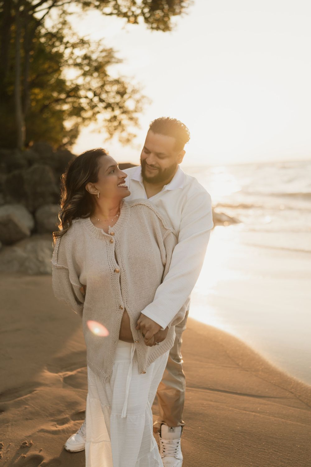 Smiling couple embracing on a sandy beach at golden hour sunset, with waves, rocks and trees in the background.