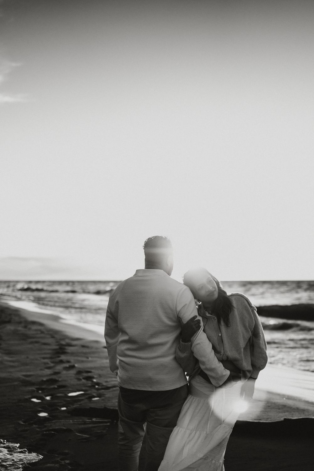 Black and white photo of a couple embracing on a beach, her head resting on his shoulder as they gaze out at the ocean waves.