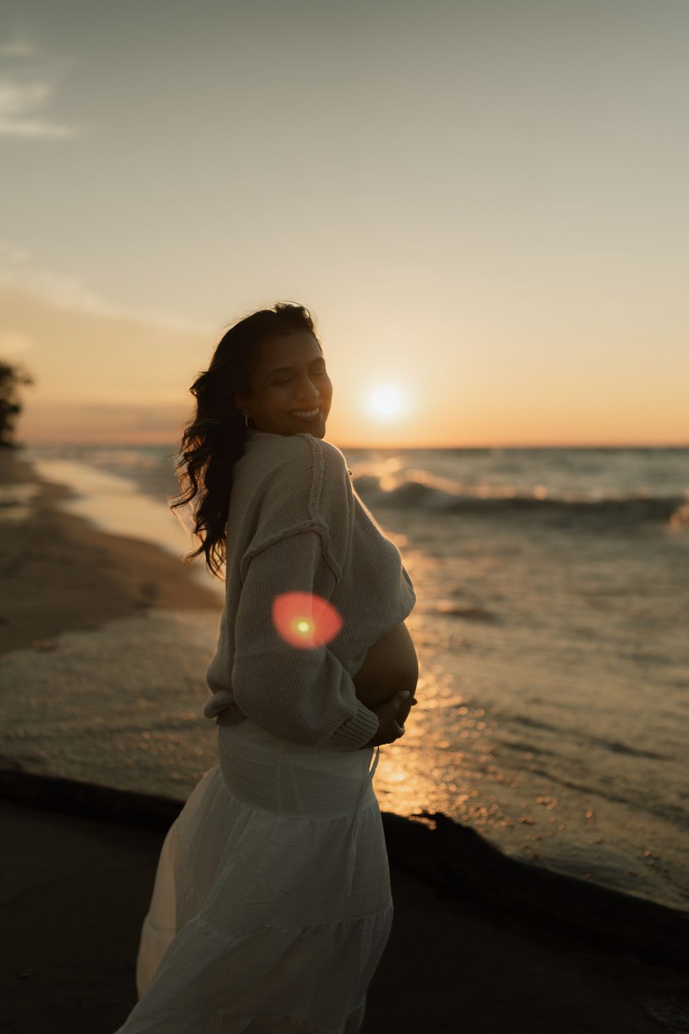 Smiling pregnant woman in a white skirt and sweater cradles her baby bump on a beach at golden sunset with waves and sun flare.